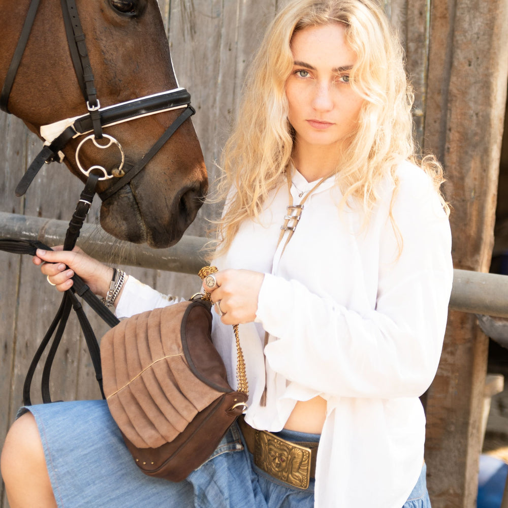 A woman in a white shirt and denim skirt sits on a brown horse, holding a brown leather shoulder bag with a gold chain strap. The background shows a rustic wooden fence and stable door.